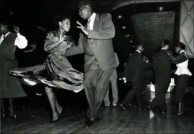 A couple swing dancing on a crowded ballroom floor, the woman's skirt flaring out